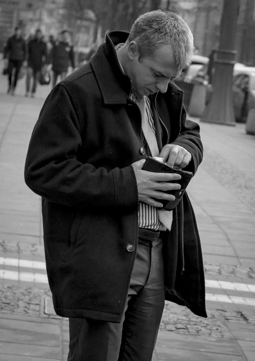 Man in a dark coat looking down into his wallet on a city sidewalk